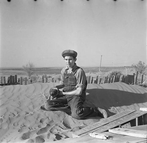 Image shows a young boy holding what appears to be a football sitting on top of a very large dune of dust.  It looks like he is sitting on a sand dune in a desert.