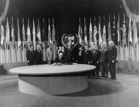 At the conference in San Francisco, Secretary of State Edward Stettinius signs the United Nations charter while President Harry S. Truman (second from left) looks on.  The United States delegation is gathered about.  