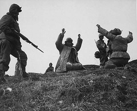 In a grassy hill area, two men are seen kneeling with their arms in the air. Three soldiers are seen on each side of them with guns.  All men are dressed in cold weather gear.