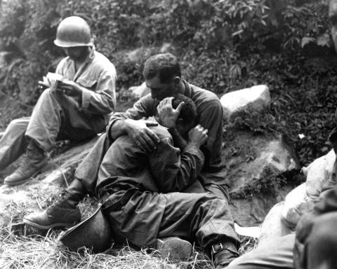 Three men are seated on a grassy area with a hill behind them.  The soldier in the background wears a helmet and is writing on a booklet of some kind.  In the center of the photo, one soldier is cradled in the arms of another soldier in the way a mother would often cradle a crying child.