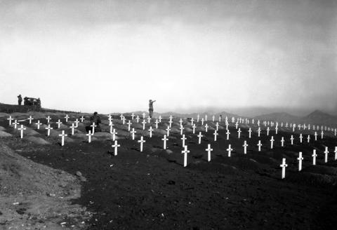 A lone bugler is seen in the background of a Marine cemetery at Hamhung, Korea in 1950.  Dozens of graves are marked with white crosses, each mounded with dirt.  Bare soil is seen throughout the cemetery - no grass has grown.  In the distance is a vehicle and three soldiers standing at attention.  One additional man is seen kneeling amid the graves.