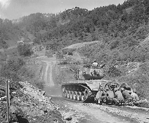 Five uniformed army soldiers are seen kneeling behind a tank and one man is seen coming out of the top of the tank.  Tank is traveling on dirt road in the rural Hongcheon area of Korea in 1951 and firing on Communist troops seen in the distance.