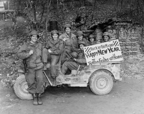 Seven uniformed, smiling, relaxed soldiers from Iowa are seen seated on an army jeep. They hold a sign that reads: “Rock of the Marne • Happy New Year • To The Folks at Home.” The background is a wooded hillside with a large amount of sandbags stacked up.