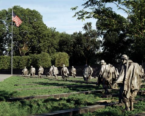 Multiple statues visible in an are of low-lying bushes and granite blocks.  United States flag is seen on the left.  Mature trees are seen in the background.