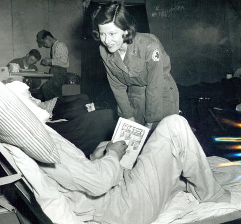 Woman identified as Red Cross worker Mary Jane White of Burlington, Iowa is seen talking with a wounded soldier at a M.A.S.H. hospital.  Ms. White wears a uniform with a Red Cross logo on the left sleeve.  She is bent down in conversation with a wounded soldier who is reclining on a hospital cot.  The patient’s head is bandaged, and he holds a magazine in his hands.