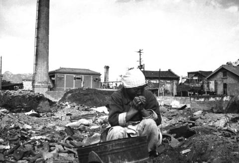 Elderly Korean woman wearing plain-styled shirt, skirt, and head-covering, sits in rubble of some sort of structure. 