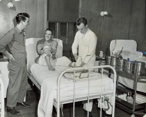 Three men seen in Great Lakes Hospital in Korea.  One man sits smiling on a hospital bed while another man bandages his left foot.  A third man is seen talking with the injured man.  Medical equipment sits on a cart near the man who is doing the bandaging.