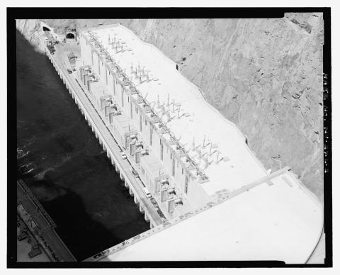 The image shows an aerial view of Hoover Dam with a power station at the top.  On one side there is visible water, and on the other there is none. 