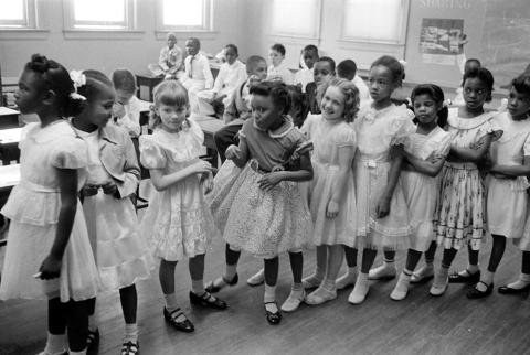 Photograph shows a line of African American and white school girls standing in a classroom while boys sit behind them.