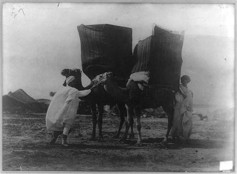 Image shows two men in the Sahara with very large wares bags on the backs of two camels. 