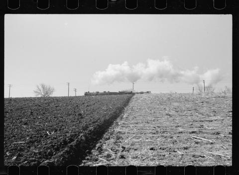 Image of Iowa land that has been freshly plowed on one side of the image, and not plowed on the other.  There is a train and telephone poles in the background.
