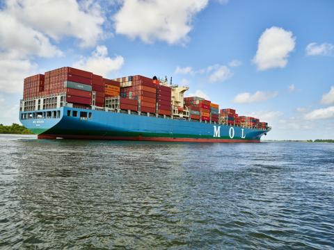 Color photo of a loaded cargo ship navigates the Savannah River.  “MOL” is visible on the side of the ship.