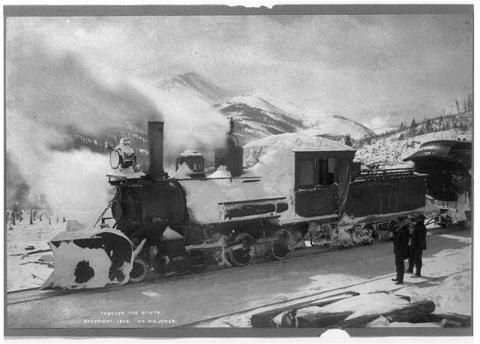 Snow-covered locomotive stopped on railroad tracks with mountains visible in the background.  Two men stand near the train in the foreground.