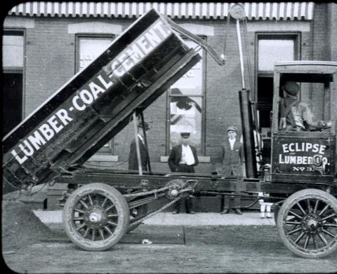 Old-fashioned delivery truck with box hoisted in the air.  “Lumber - Coal - Cement” printed on the side of the box, and four men are also seen in the photo.