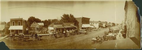Landscape photo of Main Street in Elliott, Iowa with many buildings, awnings, and horse-drawn wagons parked in front of buildings.