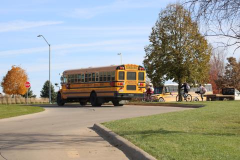 Two school busses in Pella, Iowa, 2018.  One bus is about to pull out onto the street and the other bus is about to turn into the school drive.  Two kids riding bicycles and another student standing on the sidewalk are also visible.