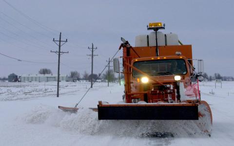 Large, orange snowplow removing snow on a paved road somewhere in rural Iowa.  The only person in the photo is the driver of the snowplow.