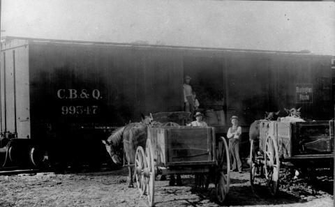Two horse-drawn wagons parked near train cars.  Doors are open on train cars, and three men and a child are also seen in the photo.