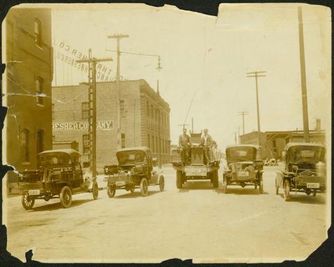 Four “Model T” roadsters and one Peerless Truck parked on a Des Moines street.  Electrical poles and brick buildings are also visible.