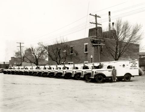 Sixteen delivery trucks lined up along with their drivers just outside of the Farmers Mutual Co-Op Creamery.  