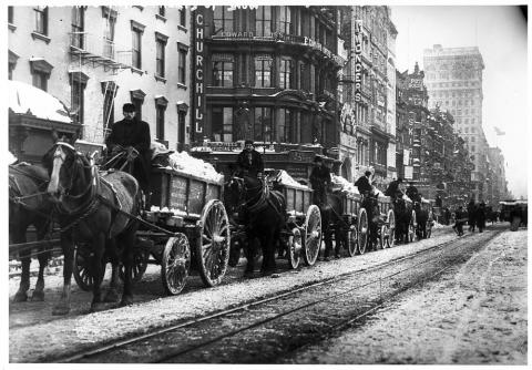 Six, horse-drawn, snow-piled wagons lined up outside of brick buildings and next to streetcar tracks in New York City.