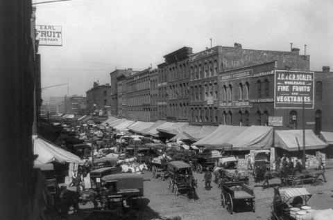 Black-and-white photo of many horse-drawn wagons moving in and out of wagon-lined street.  Multi-story buildings have very large awnings on front.