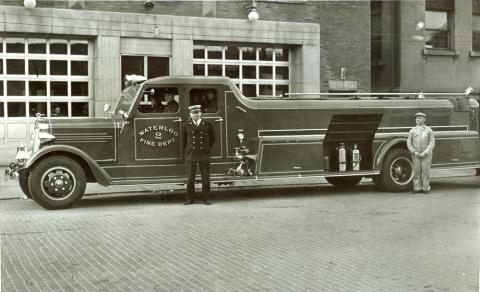 Waterloo Fire Department displays their new fully enclosed fire truck.  The new truck is parked outside the fire station, along with two men standing near it.