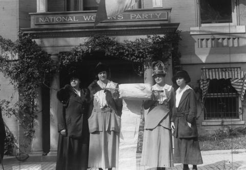 Four women in dresses and hats proudly stand in front of a building with a sign that reads “National Women’s Party.”  Between the two women in the center is a very large roll of paper that the women are holding up.  These National Women’s Party leaders had been working to collect signatures of support on a “mile long” paper to present to members of Congress.