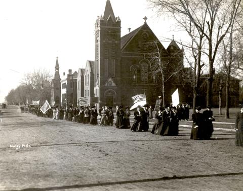 In this photograph, taken in Boone, Iowa on October 29, 1908, a suffrage parade made up of people carrying banners and flags passes by a large church. One banner reads: “TAXATION WITHOUT REPRESENTATION IS TYRANNY. AS TRUE NOW AS IN 1776.” 