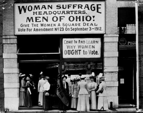 In this 1912 picture taken outside the Woman Suffrage Headquarters in Upper Euclid Avenue in Cleveland, Ohio, four suffragettes are seen conversing outside the building (including Belle Sherman, President of the National League of Women Voters), six are doing the same inside the doorway, and one man, Judge Florence E. Allen, is standing on the sidewalk holding a flag stating, “VOTES FOR WOMEN.”