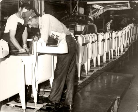 Two adult men in foreground bent over working on washing machines that are closely lined up on an assembly line.  Two women and a man are seen in the background also  working on a part of the Maytag washing machine.