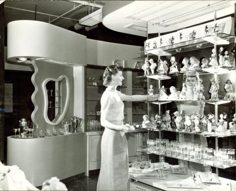 Adult woman standing in front of six-shelf display of glassware in J.C. Penny department store.  