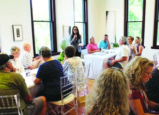 A person holding a microphone addresses a crowd of people seated at tables.