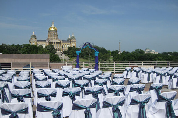 Many rows of chairs with decorative covers set up for a wedding ceremony on the Cafe Terrace of the State Historical Building with a view of the Iowa State Capitol building.