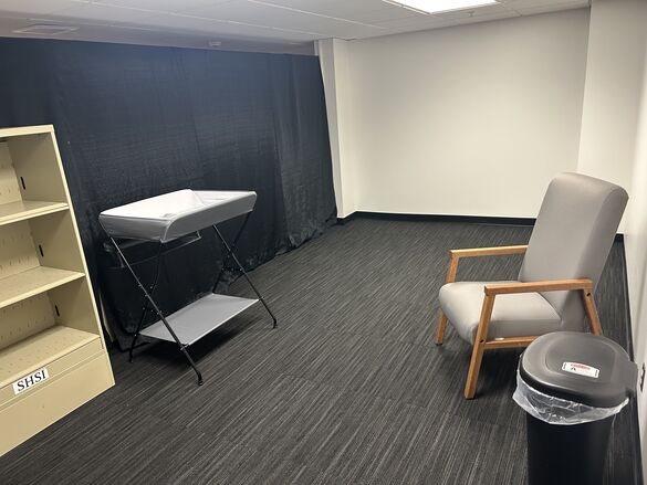 Lactation space for visitors with a shelf, baby changing table, chair, and trash can.