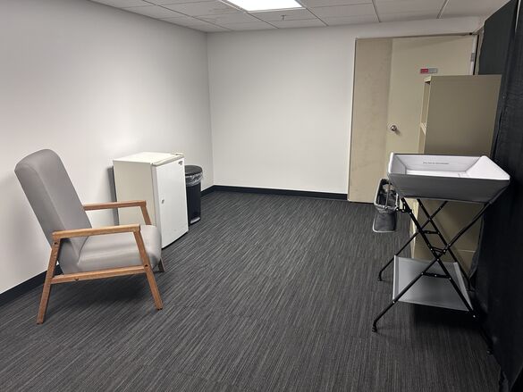 Lactation space for visitors with a chair, mini fridge, shelf, baby changing table, and trash can.