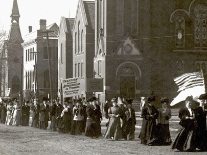 Women marching, holding signs and U.S. flags as part of the Women's Suffrage March in Boone, IA in 1908.